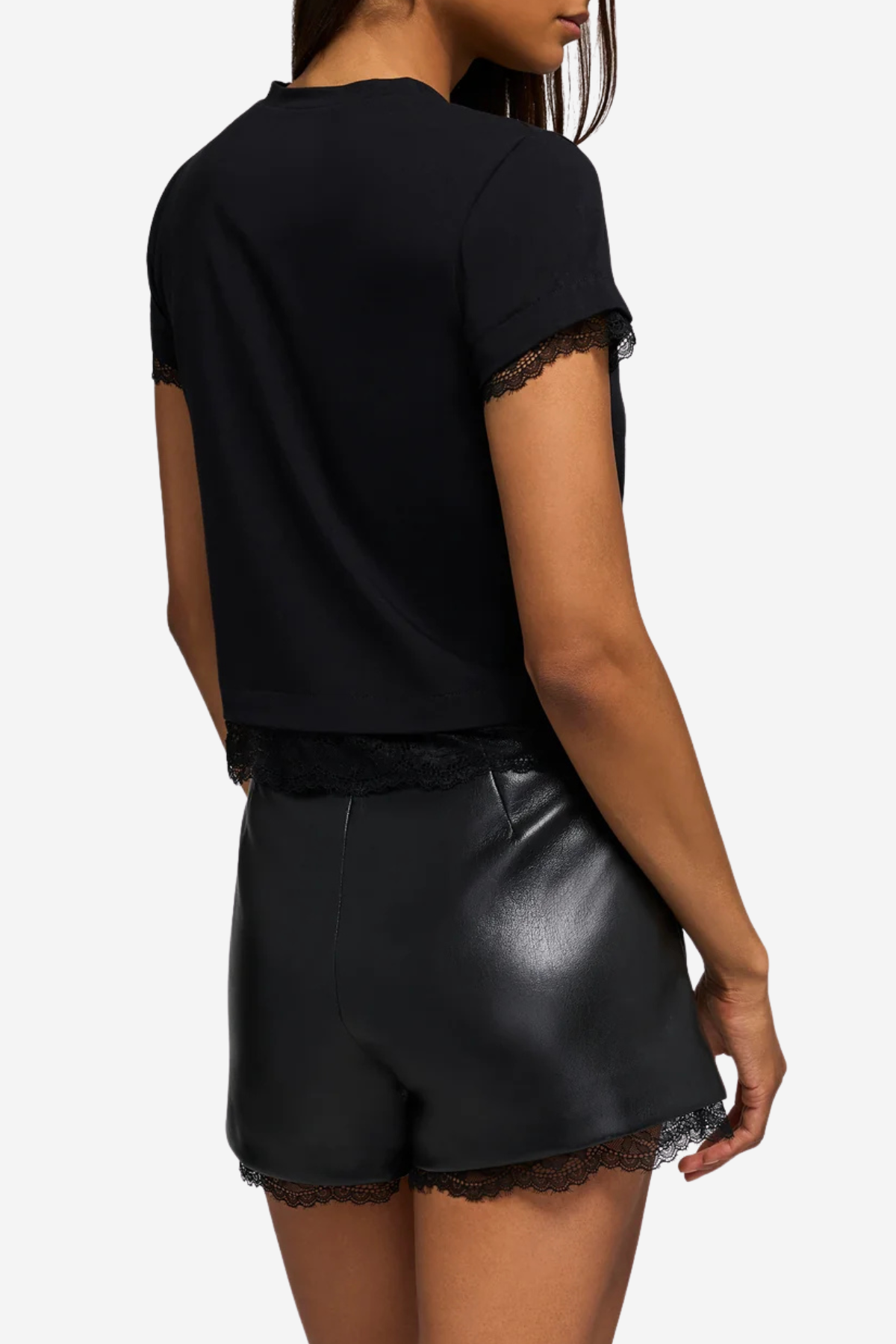 A woman shown from the back wearing a black short-sleeve top with lace trim at the hem and sleeves, paired with black faux-leather shorts with lace trim, against a light gray studio background.