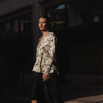 A woman walking outdoors at night wearing a cream blouse with a black monogram pattern and a black midi skirt.