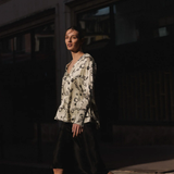 A woman walking outdoors at night wearing a cream blouse with a black monogram pattern and a black midi skirt.