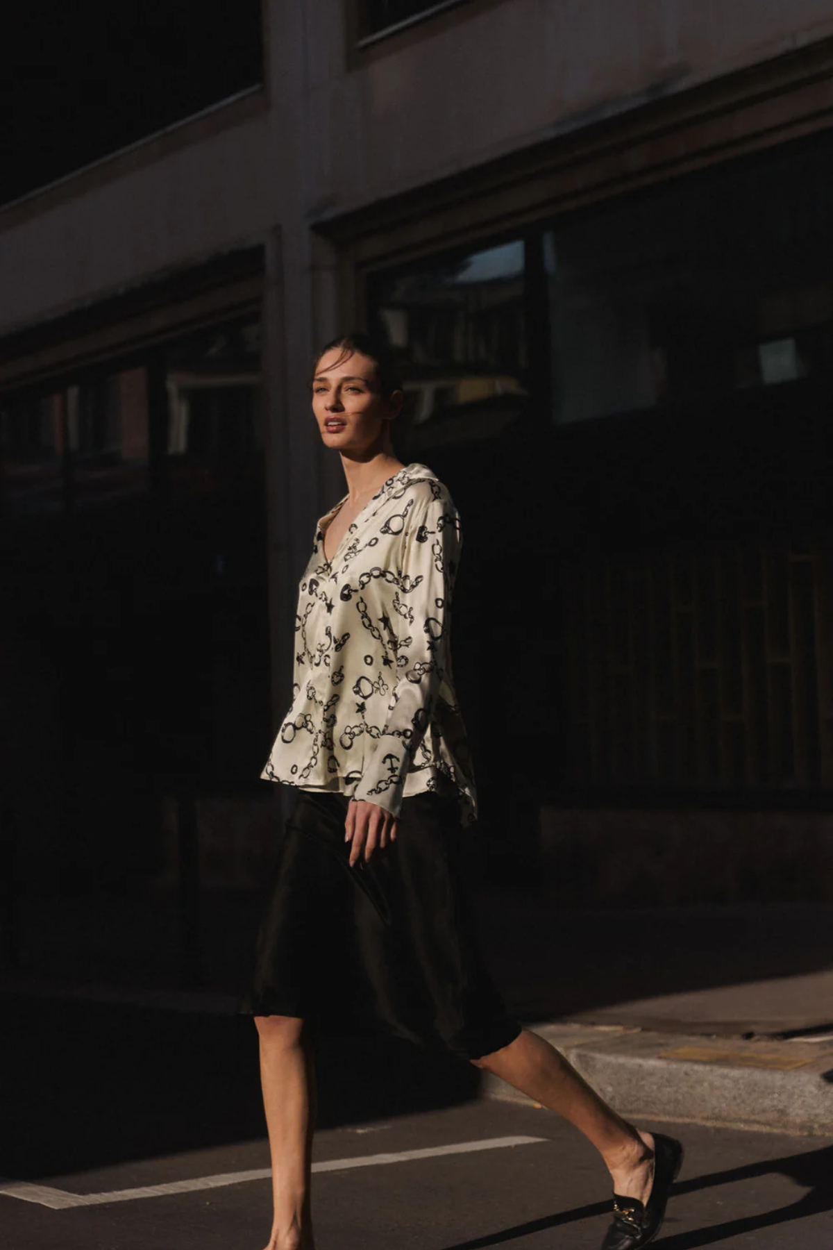 A woman walking outdoors at night wearing a cream blouse with a black monogram pattern and a black midi skirt.