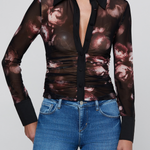 A woman wearing a sheer long-sleeve button-front blouse with a dark floral print and black collar, cuffs, and placket, paired with blue jeans against a light gray studio background.