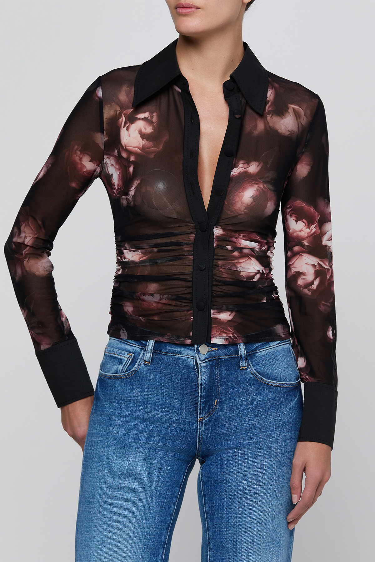 A woman wearing a sheer long-sleeve button-front blouse with a dark floral print and black collar, cuffs, and placket, paired with blue jeans against a light gray studio background.