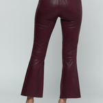 A person shown from the back wearing high-waisted, flared trousers in a deep burgundy faux-leather material, paired with a matching long-sleeve top and pointed-toe heels, against a light gray studio background.