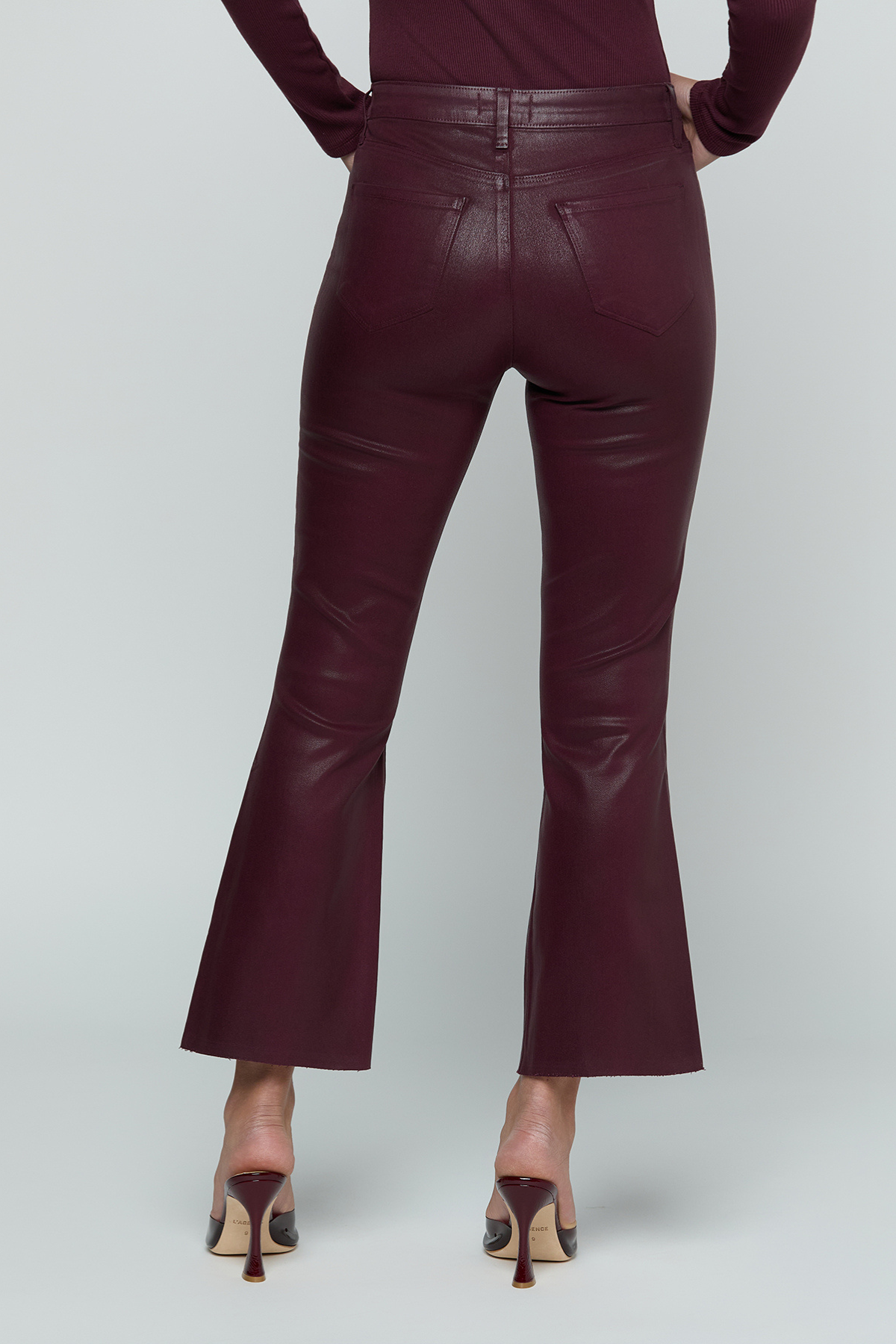 A person shown from the back wearing high-waisted, flared trousers in a deep burgundy faux-leather material, paired with a matching long-sleeve top and pointed-toe heels, against a light gray studio background.