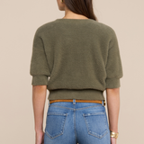 Model wearing an olive green sweater with jeans and a belt on a beige background.