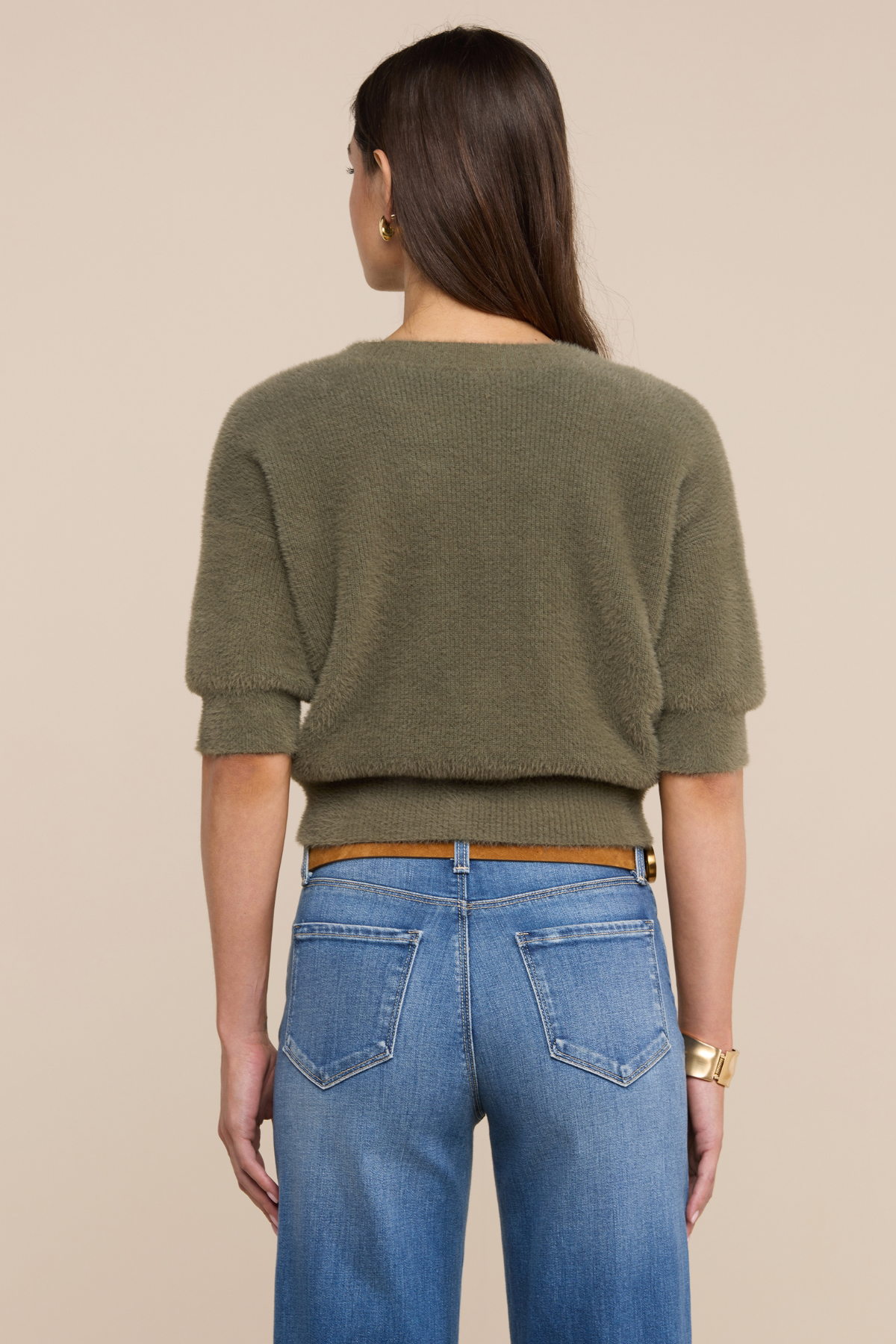 Model wearing an olive green sweater with jeans and a belt on a beige background.