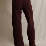 A person wearing loose, straight-leg knit pants in a deep burgundy or maroon shade, shown from the waist down against a draped off-white fabric backdrop.