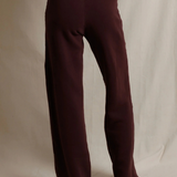 A person wearing loose, straight-leg knit pants in a deep burgundy or maroon shade, shown from the waist down against a draped off-white fabric backdrop.