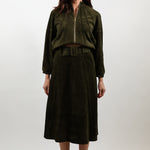 A woman stands against a light gray studio background wearing a dark olive green two-piece outfit consisting of a cropped zip-front jacket and a matching midi skirt with a belt, paired with light beige pointed heels.