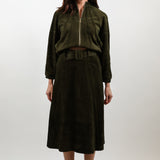 A woman stands against a light gray studio background wearing a dark olive green two-piece outfit consisting of a cropped zip-front jacket and a matching midi skirt with a belt, paired with light beige pointed heels.
