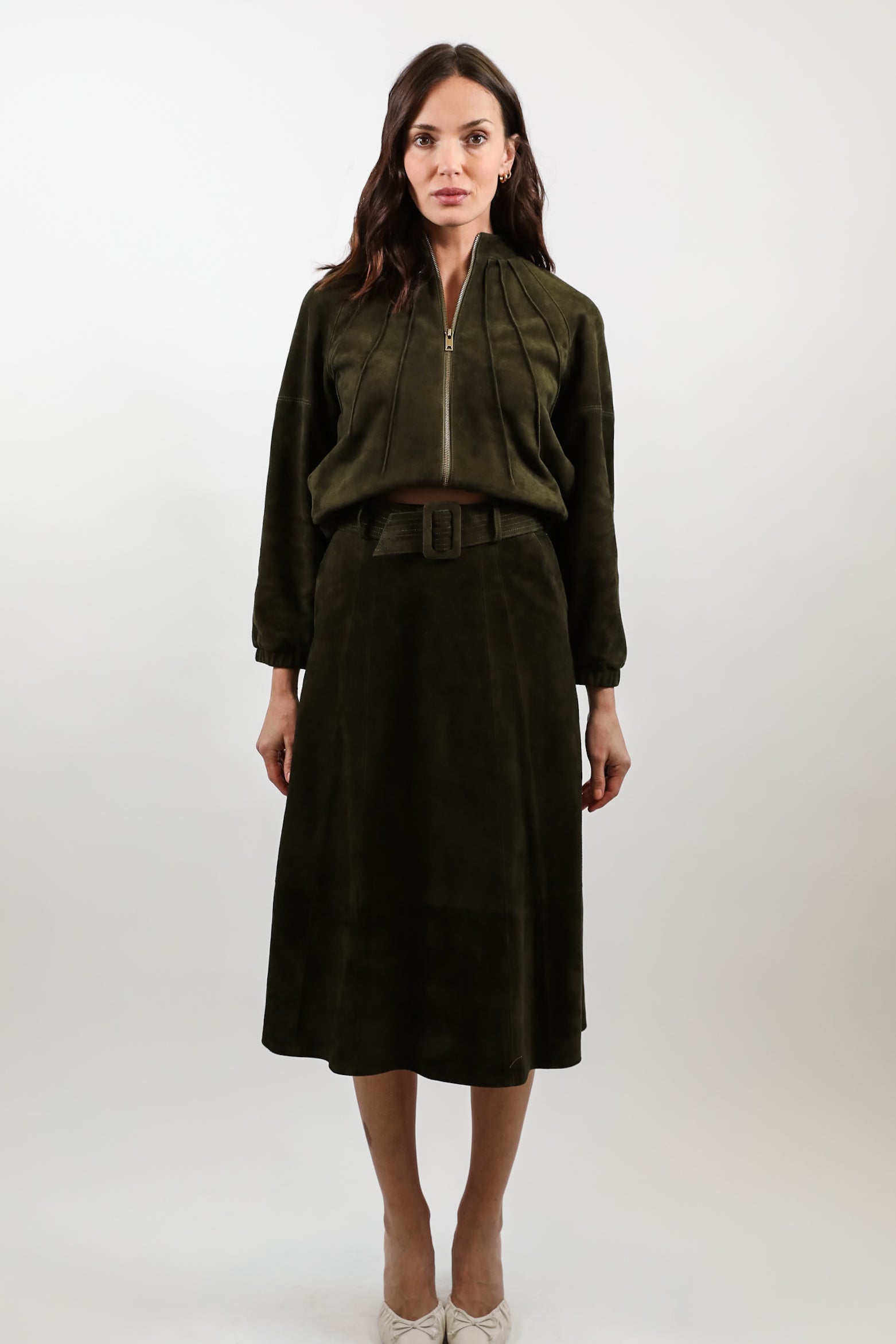A woman stands against a light gray studio background wearing a dark olive green two-piece outfit consisting of a cropped zip-front jacket and a matching midi skirt with a belt, paired with light beige pointed heels.