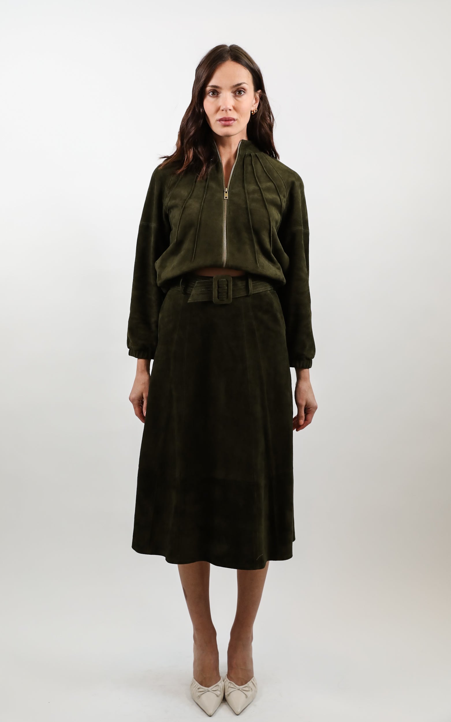A woman stands against a light gray studio background wearing a dark olive green two-piece outfit consisting of a cropped zip-front jacket and a matching midi skirt with a belt, paired with light beige pointed heels.