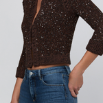 A woman wearing a dark brown sparkly knit cardigan with three-quarter sleeves and blue jeans, shown from the neck down against a light gray background.