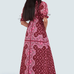 A woman modeling a long, dark red maxi dress with pink and light pink paisley patterns, shown from the back against a light gray studio background.