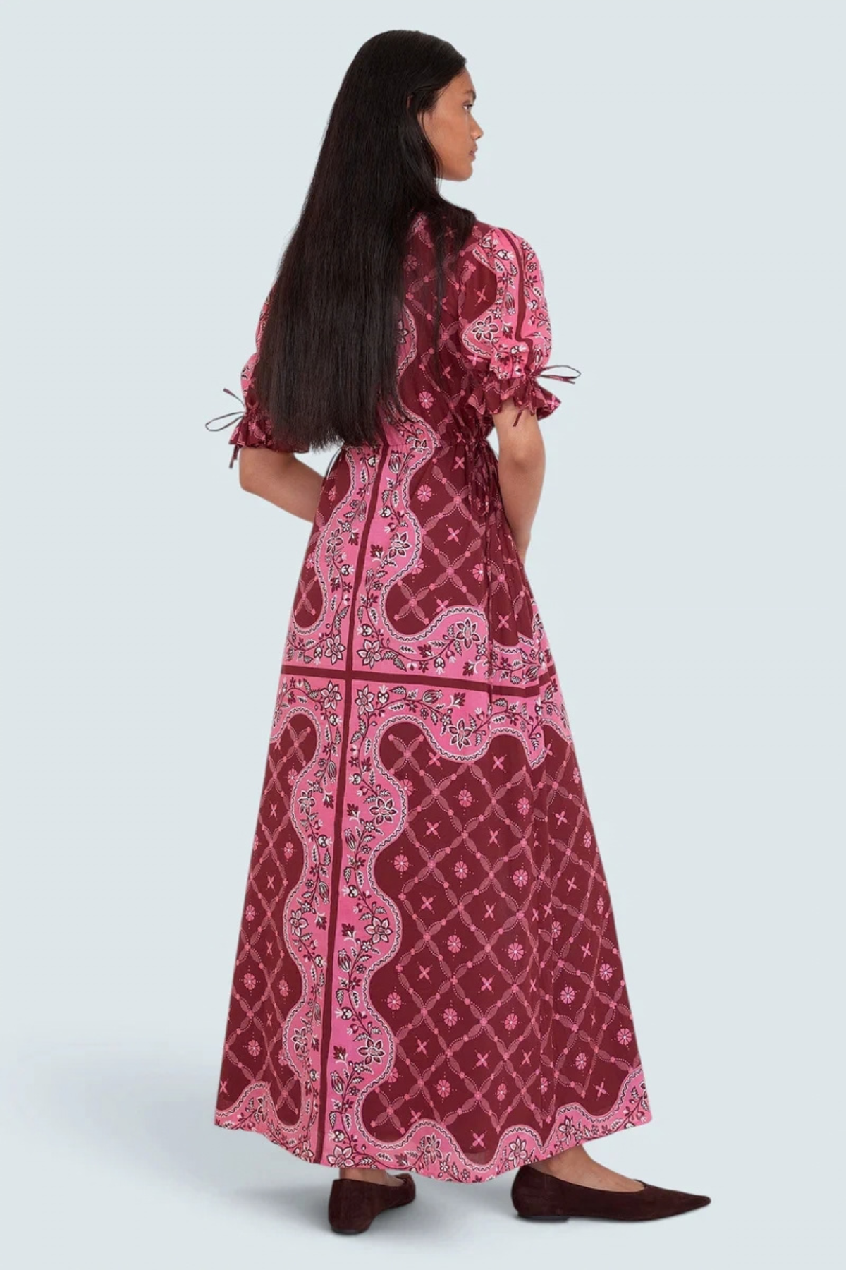 A woman modeling a long, dark red maxi dress with pink and light pink paisley patterns, shown from the back against a light gray studio background.