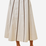 An off-white A-line midi skirt with vertical dark brown pleats, shown on a model wearing clear high-heeled sandals against a light gray background.