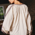 A woman wearing a cream-colored, oversized long-sleeve blouse with gathered detailing and embroidered trim on the shoulders and cuffs, paired with olive green pants, shown from a side-back angle in a rustic indoor setting.