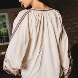 A woman wearing a cream-colored, oversized long-sleeve blouse with gathered detailing and embroidered trim on the shoulders and cuffs, paired with olive green pants, shown from a side-back angle in a rustic indoor setting.