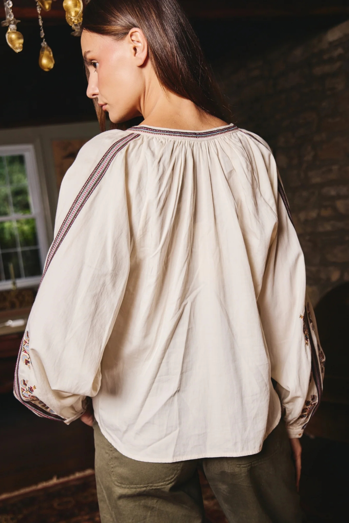 A woman wearing a cream-colored, oversized long-sleeve blouse with gathered detailing and embroidered trim on the shoulders and cuffs, paired with olive green pants, shown from a side-back angle in a rustic indoor setting.