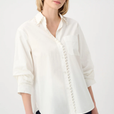A woman modeling a white long-sleeve button-up shirt with decorative button details down the front and a chest pocket, paired with dark pants against a light gray studio background.