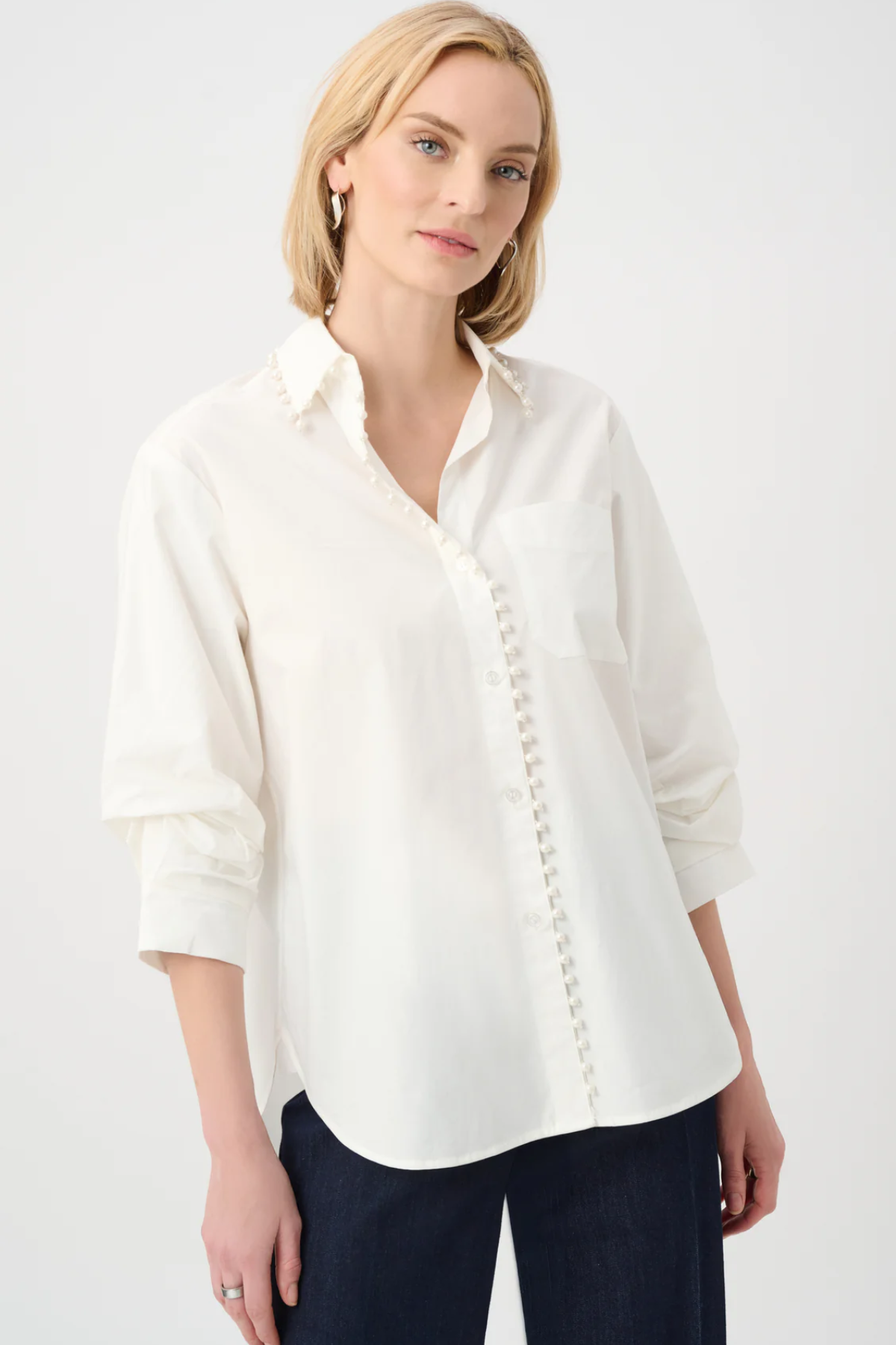 A woman modeling a white long-sleeve button-up shirt with decorative button details down the front and a chest pocket, paired with dark pants against a light gray studio background.