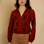 Woman wearing a red blouse with black floral patterns and brown pants against a beige background