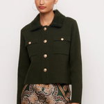 A woman modeling a dark olive green cropped jacket with a fuzzy collar and gold-tone buttons, paired with a patterned skirt, against a plain light gray background.