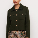 A woman modeling a dark olive green cropped jacket with a fuzzy collar and gold-tone buttons, paired with a patterned skirt, against a plain light gray background.