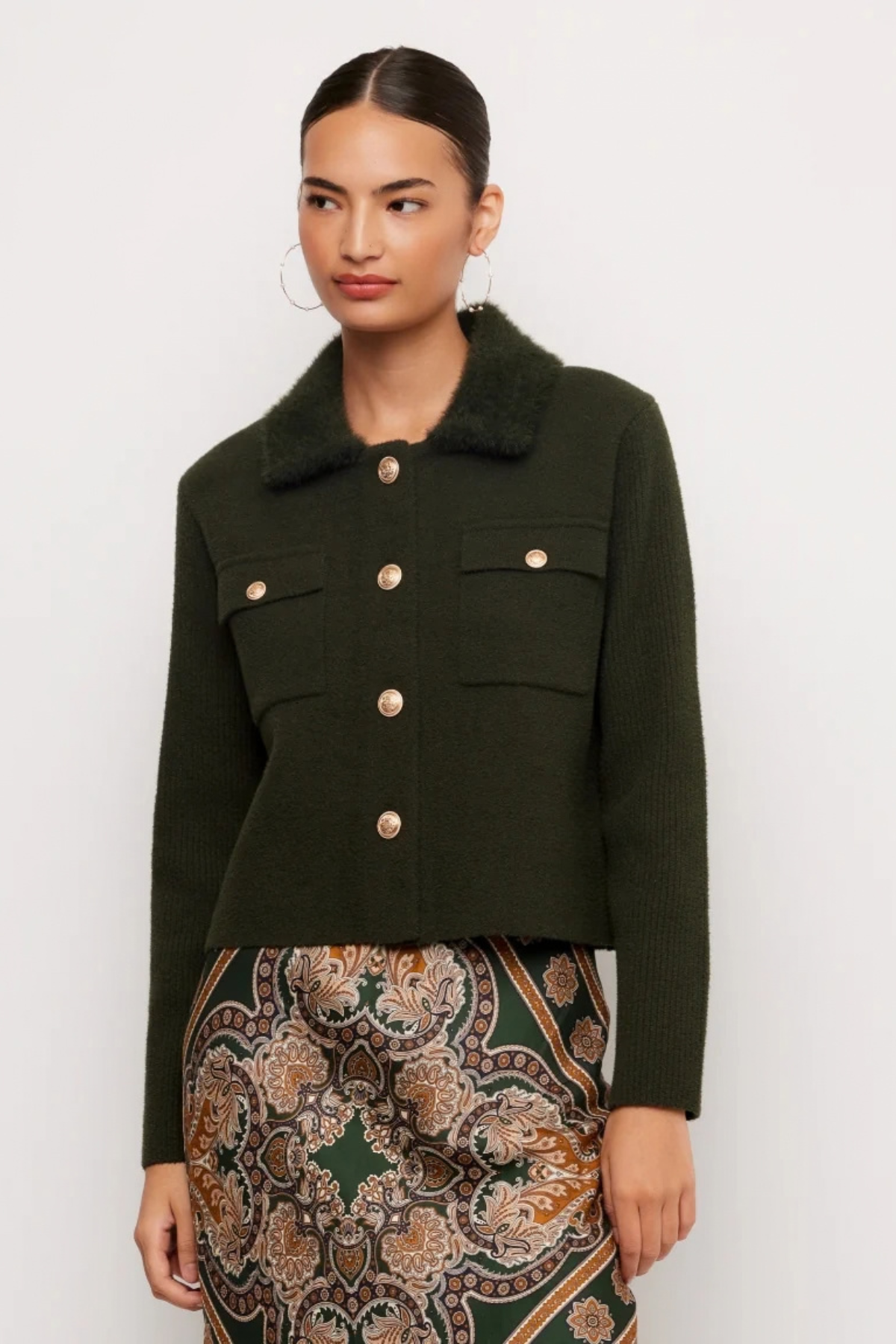 A woman modeling a dark olive green cropped jacket with a fuzzy collar and gold-tone buttons, paired with a patterned skirt, against a plain light gray background.