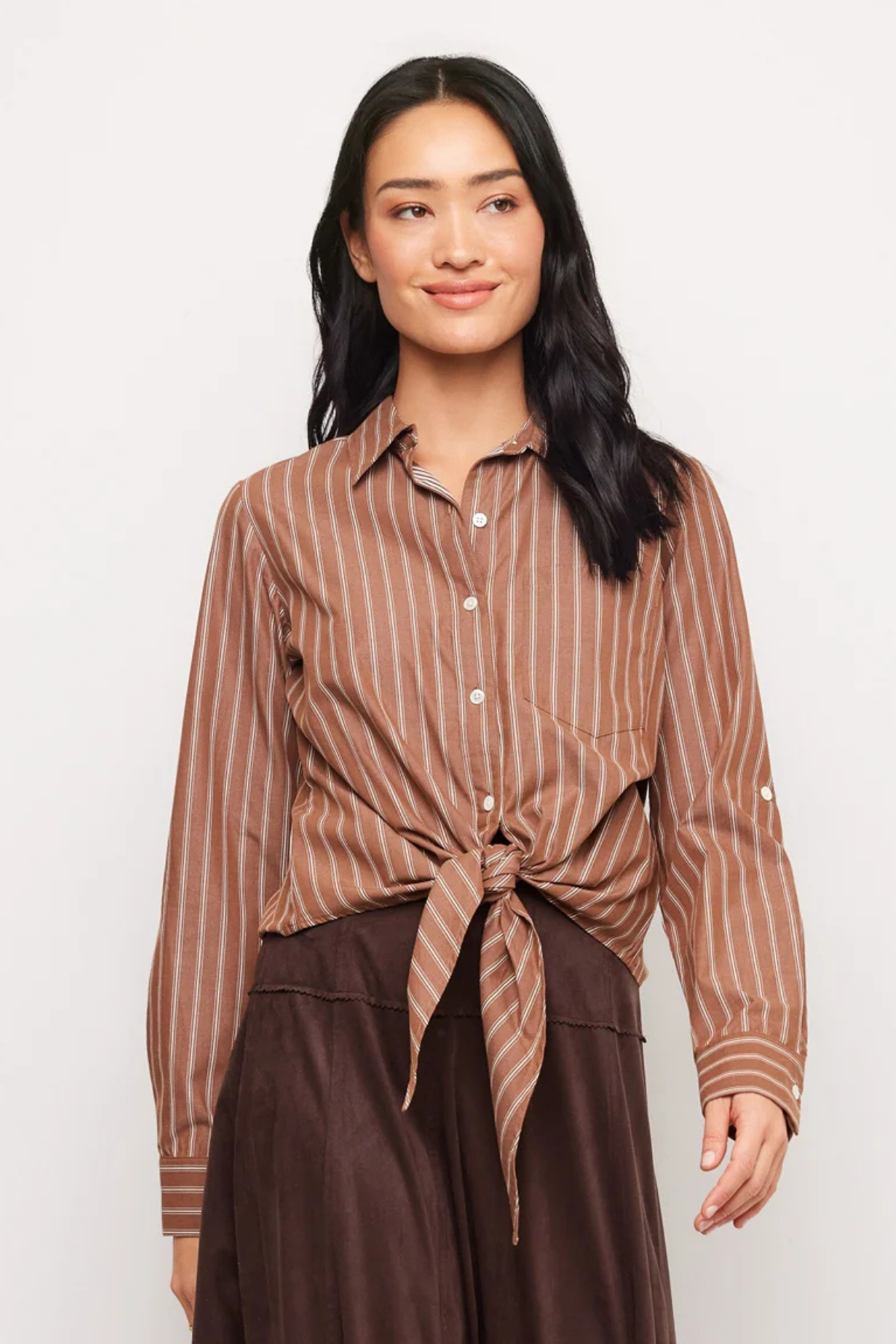 A woman modeling a brown and beige vertically striped button-down shirt tied at the waist, paired with dark brown pants, against a plain light gray background.