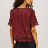 A woman shown from the back wearing a burgundy short-sleeve blouse with lace trim at the cuffs and hem, paired with black pants, against a light gray studio background.