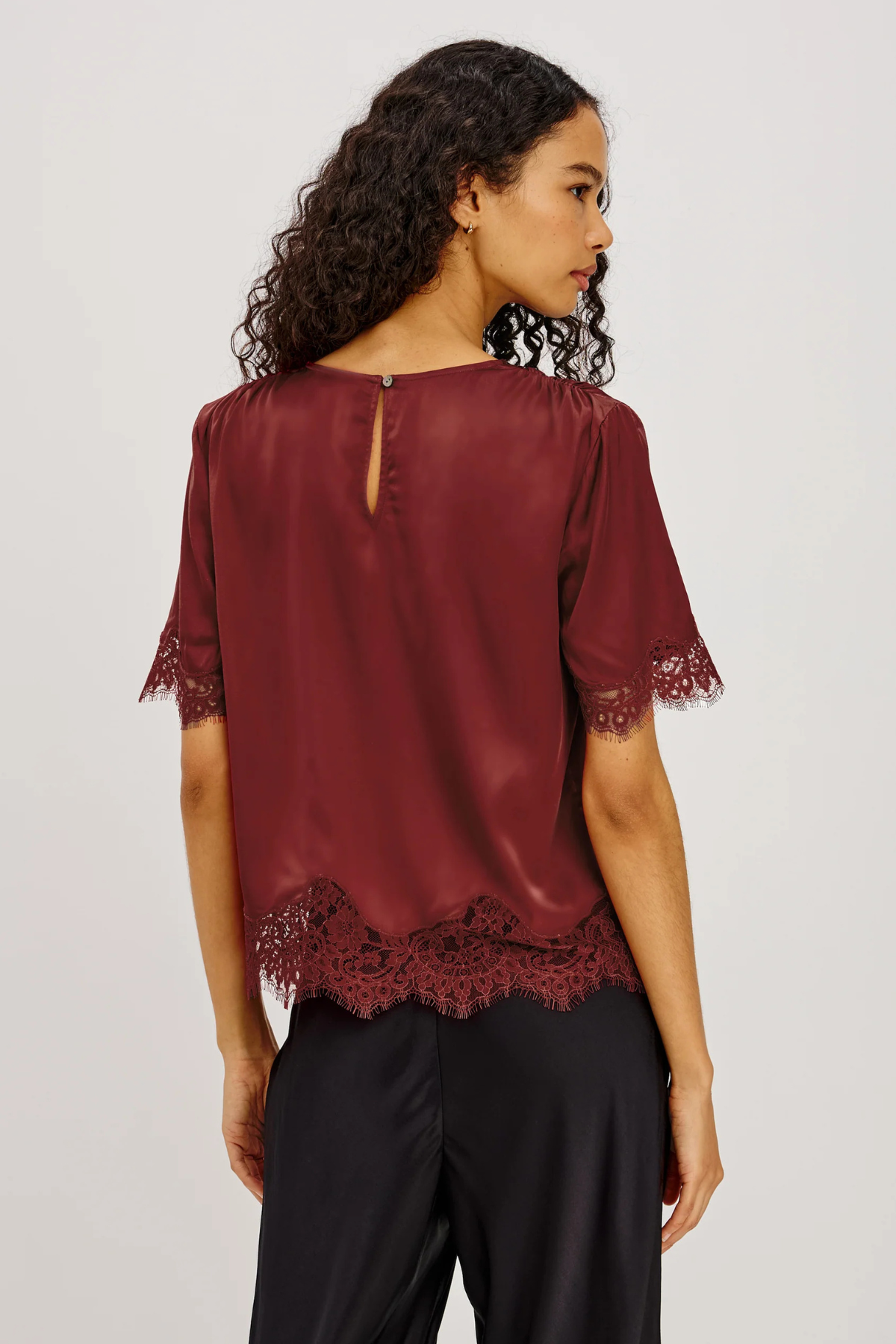 A woman shown from the back wearing a burgundy short-sleeve blouse with lace trim at the cuffs and hem, paired with black pants, against a light gray studio background.