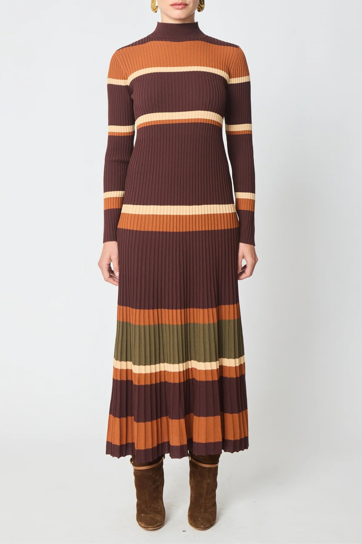 A woman modeling a long-sleeve ribbed knit mock-neck midi dress with horizontal stripes in dark brown, rust orange, olive green, and cream, paired with brown suede ankle boots against a light gray studio background.