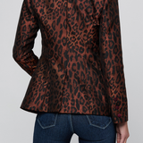 A woman shown from the back wearing a brown and black leopard-print blazer with dark red piping on the collar and cuffs, paired with dark blue jeans, against a light gray studio background.