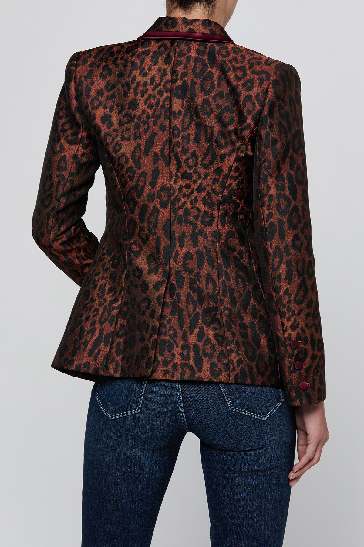A woman shown from the back wearing a brown and black leopard-print blazer with dark red piping on the collar and cuffs, paired with dark blue jeans, against a light gray studio background.