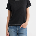 A woman wearing a black short-sleeve crewneck t-shirt with subtle white contrast stitching, paired with blue jeans, against a plain white studio background.