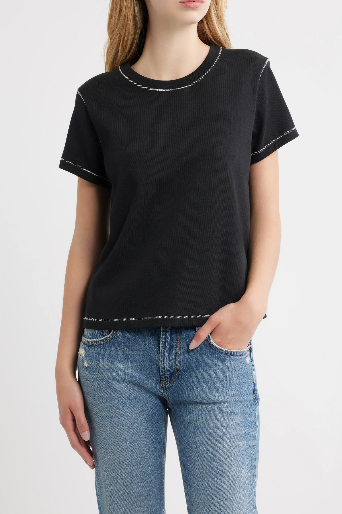 A woman wearing a black short-sleeve crewneck t-shirt with subtle white contrast stitching, paired with blue jeans, against a plain white studio background.
