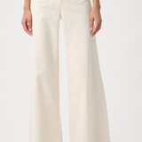 A model wearing a pair of off-white wide-leg jeans with gold-tone hardware on the front pockets, paired with beige flats, against a plain white studio background.