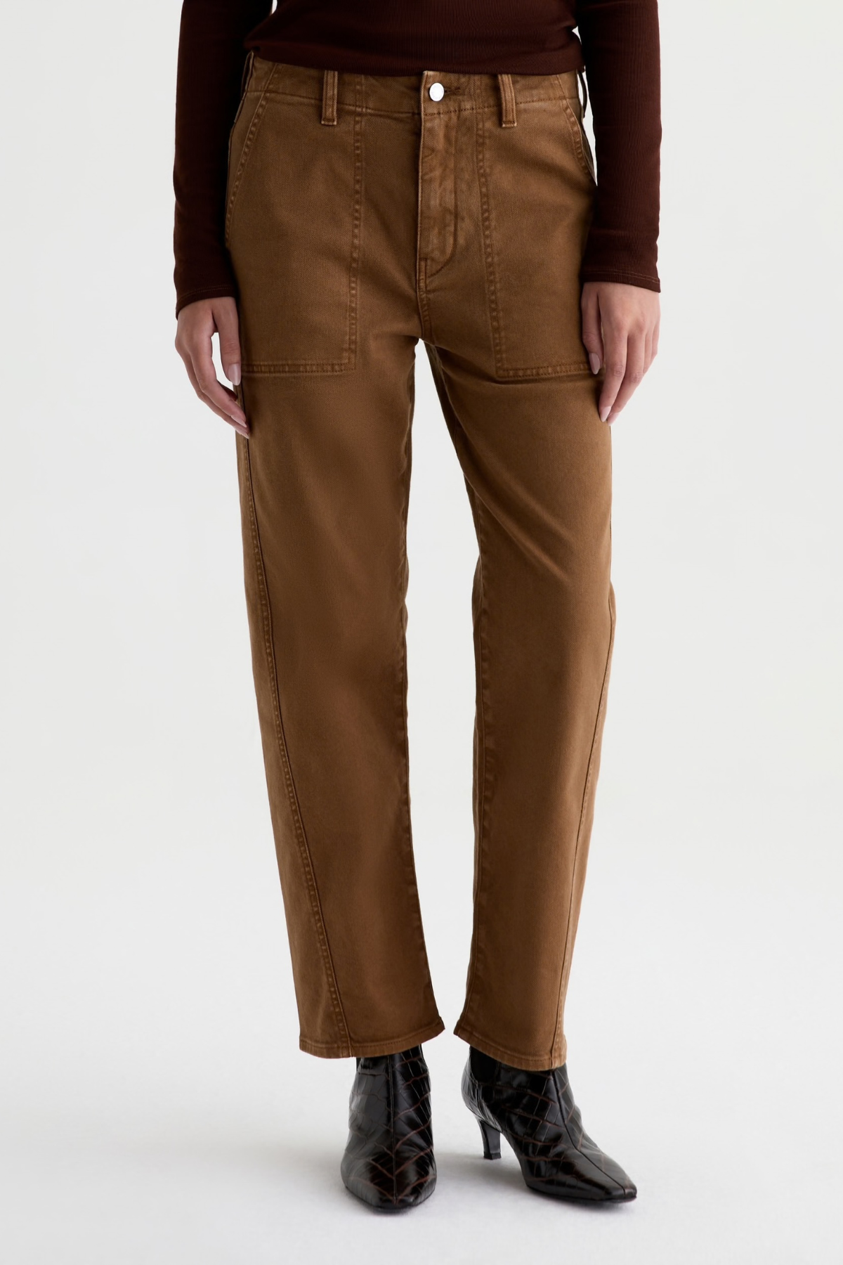 A person wearing brown straight-leg pants with a dark brown long-sleeve top and black ankle boots, shown from the waist down against a light gray studio background.