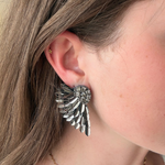 A close-up of a person's ear wearing a large silver angel wing stud earring, with brown hair and a delicate gold chain necklace visible.