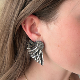 A close-up of a person's ear wearing a large silver angel wing stud earring, with brown hair and a delicate gold chain necklace visible.