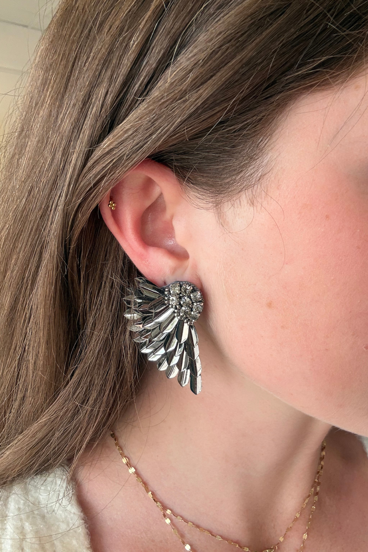 A close-up of a person's ear wearing a large silver angel wing stud earring, with brown hair and a delicate gold chain necklace visible.