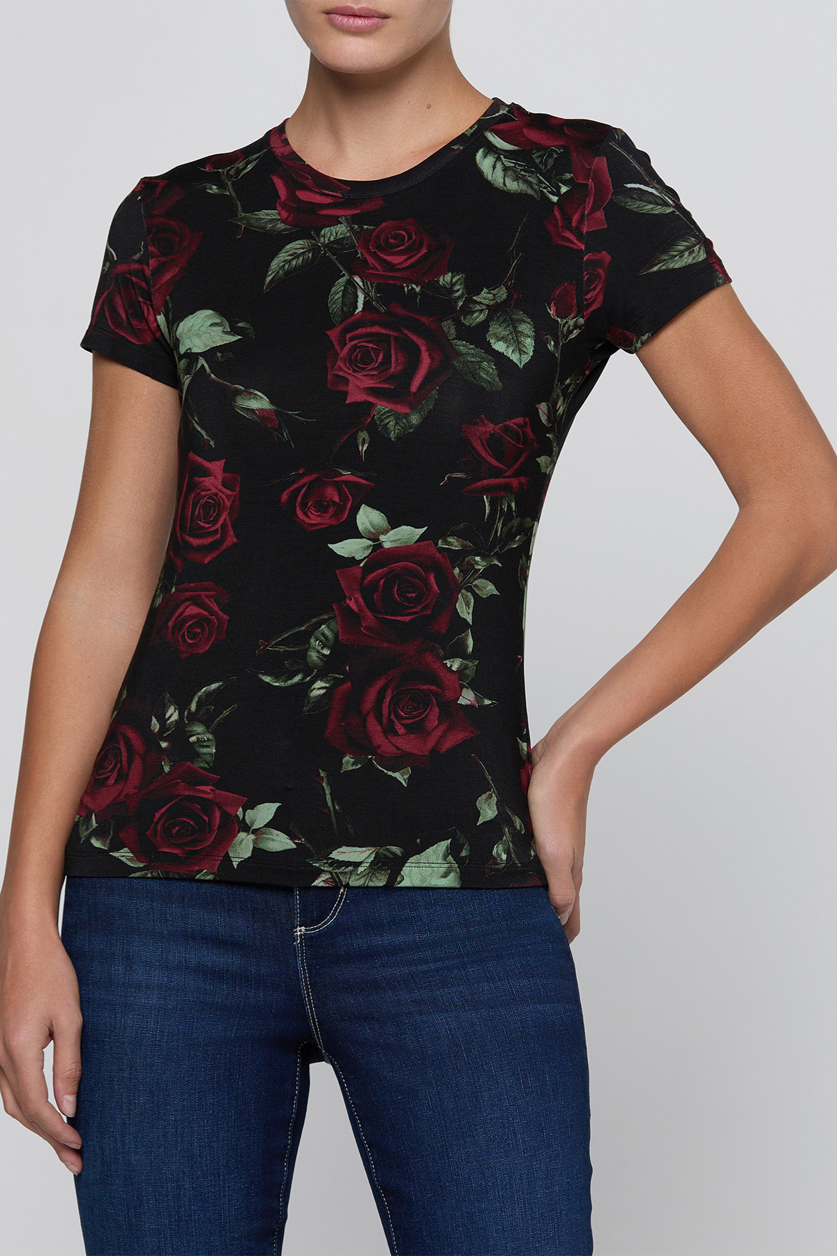 A woman wearing a black short-sleeve crewneck t-shirt with a red rose and green leaf floral print, paired with blue jeans, against a solid light gray background.