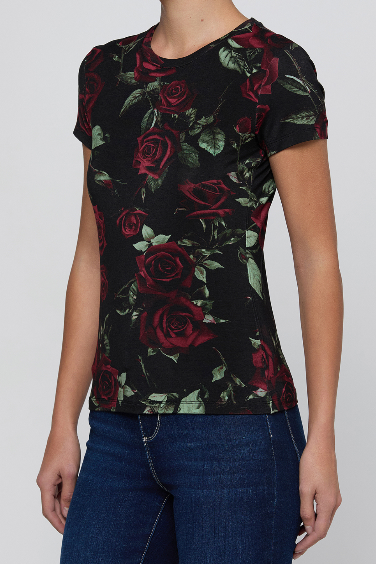 A woman wearing a black short-sleeve crewneck t-shirt with a red rose and green leaf pattern, paired with dark blue jeans, shown against a plain light gray background.