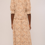 A woman viewed from the back wearing a beige and brown patterned two-piece set consisting of a short-sleeve top with ruffled shoulders and a matching long skirt, against a plain light background.