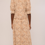 A woman viewed from the back wearing a beige and brown patterned two-piece set consisting of a short-sleeve top with ruffled shoulders and a matching long skirt, against a plain light background.