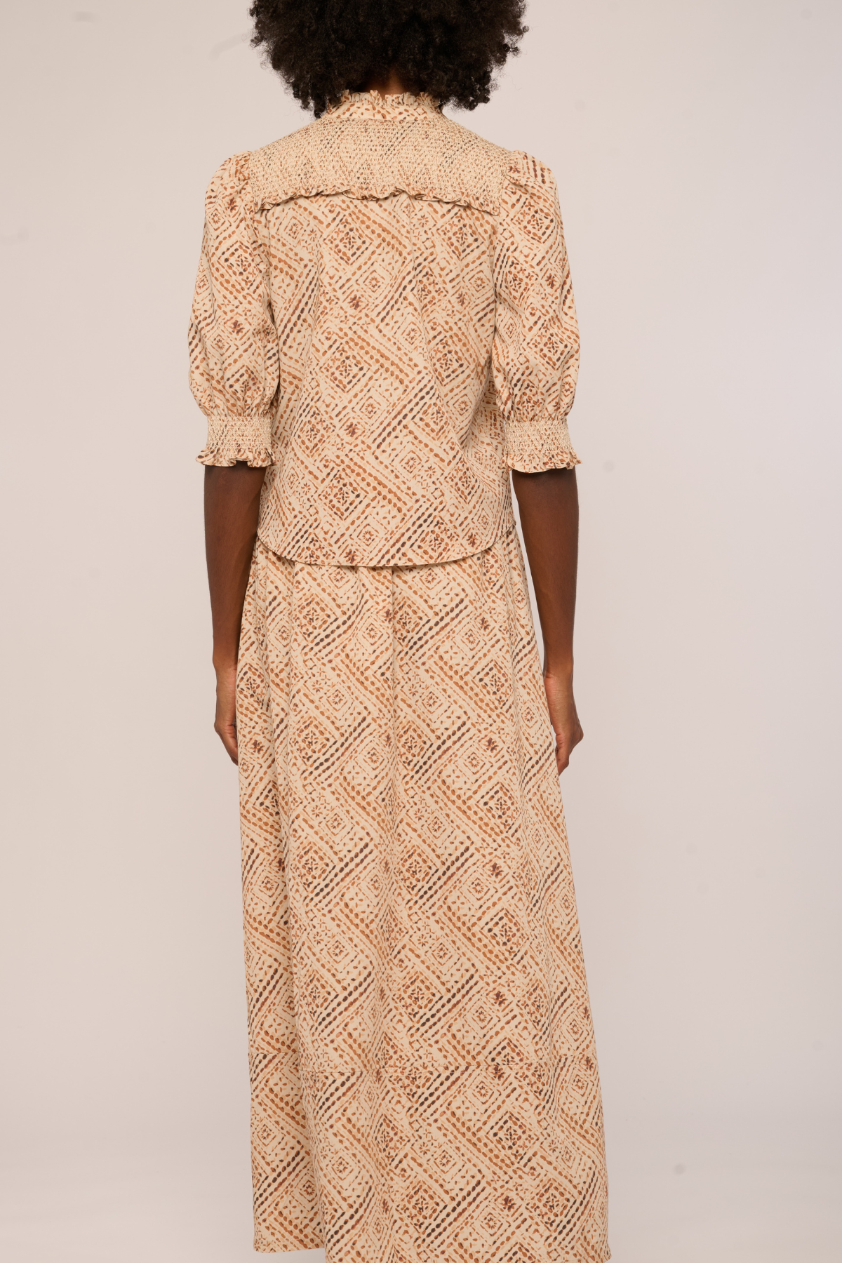 A woman viewed from the back wearing a beige and brown patterned two-piece set consisting of a short-sleeve top with ruffled shoulders and a matching long skirt, against a plain light background.