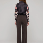 A model viewed from the back wearing a dark satin vest, a floral long-sleeve blouse, and dark brown wide-leg trousers, set against a plain light gray studio background.