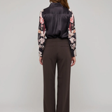 A model viewed from the back wearing a dark satin vest, a floral long-sleeve blouse, and dark brown wide-leg trousers, set against a plain light gray studio background.