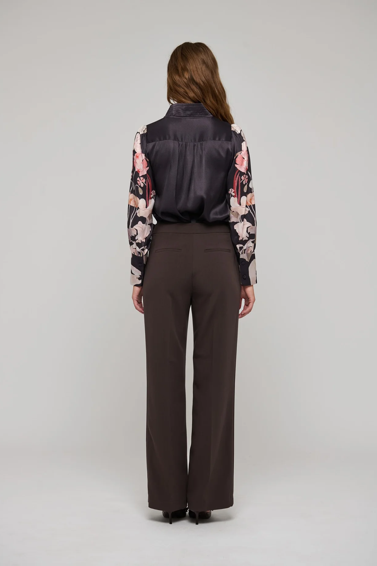 A model viewed from the back wearing a dark satin vest, a floral long-sleeve blouse, and dark brown wide-leg trousers, set against a plain light gray studio background.